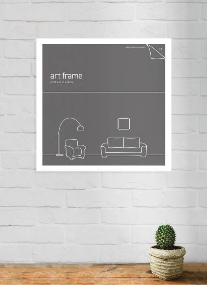A minimalist art print in a Blue Dog Posters Classic Frame 60x60cm White against a white brick wall displays a simple sketch of a living room with a sofa, armchair, floor lamp, and picture. A small potted cactus is placed on a wooden surface in the foreground. The frame size is 60cm x 60cm.