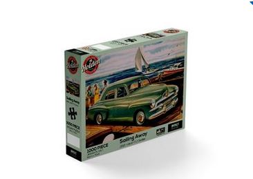 The image features a jigsaw puzzle box from Blue Dog Posters, showcasing an illustrated vintage green car near a seaside setting with a sailboat in the background. This 1000-piece puzzle is titled "Holden - Sailing Away 1000pc Puzzle.