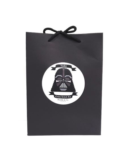 A black gift bag with a rounded sticker on the front. The sticker features a cross-stitch design of a character in a mask with the text "Hustle and Co. Cross Stitch - Vader" by Blue Dog Posters. Complete with an embroidery hoop and cross stitch fabric, the bag is elegantly tied at the top with black strings.