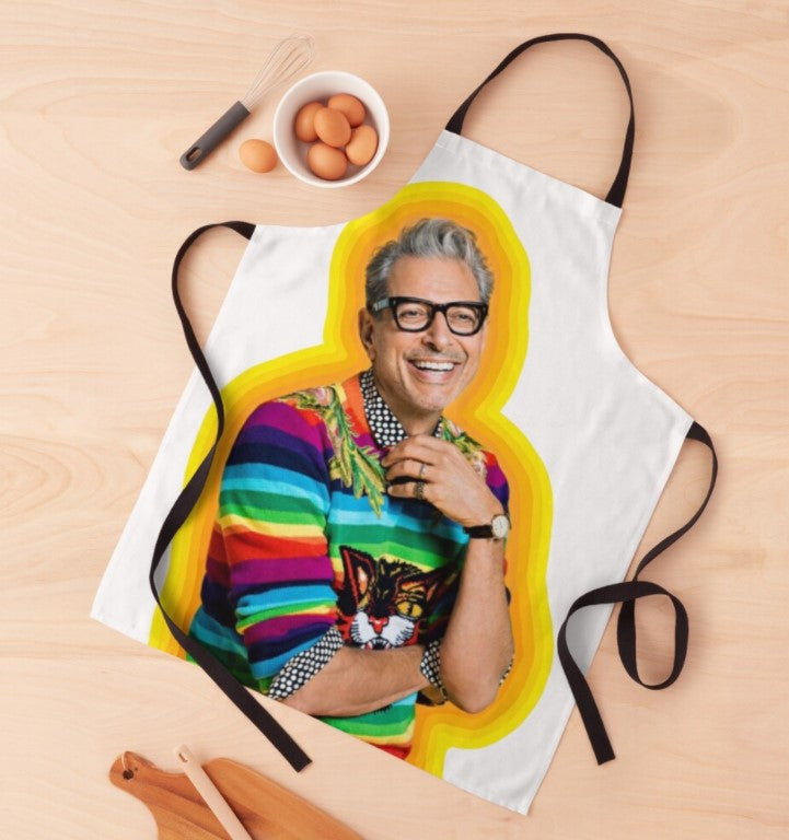 A brightly colored, machine-washable kitchen apron with a photo of a smiling person wearing glasses and a vibrant striped sweater, featuring a cat graphic. The Jeff Goldblum of Happiness Apron by Blue Dog Posters is sublimation-printed and displayed on a wooden surface with eggs in a bowl, a whisk, and wooden utensils nearby.