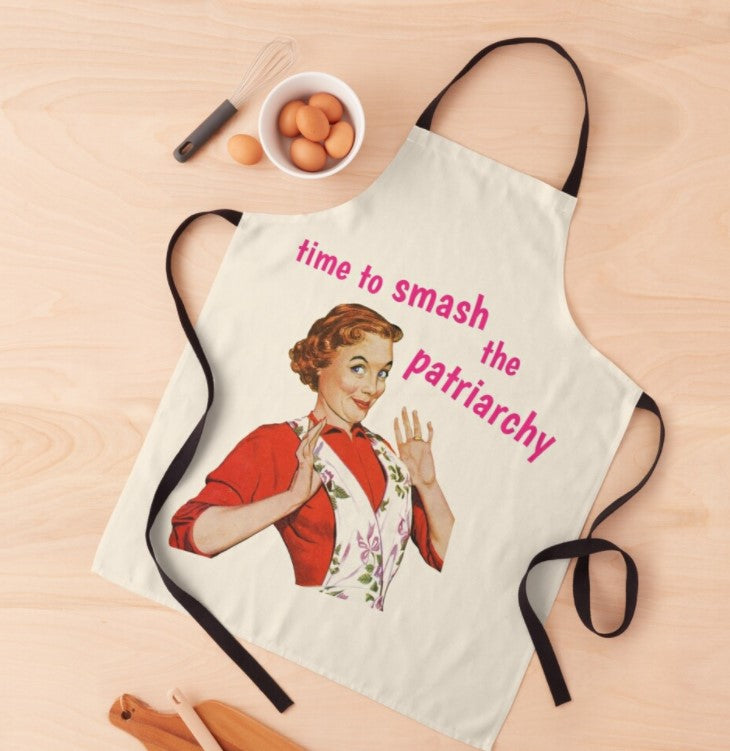 A light-colored apron with black straps features a vintage-style illustration of a woman in red, holding up her hands playfully. The text on the apron reads, "time to smash the patriarchy" in bold pink letters. Part of our Homewares Collection, this machine washable Time to Smash the Patriarchy Apron by Blue Dog Posters is displayed on a wooden surface with kitchen utensils and eggs nearby.