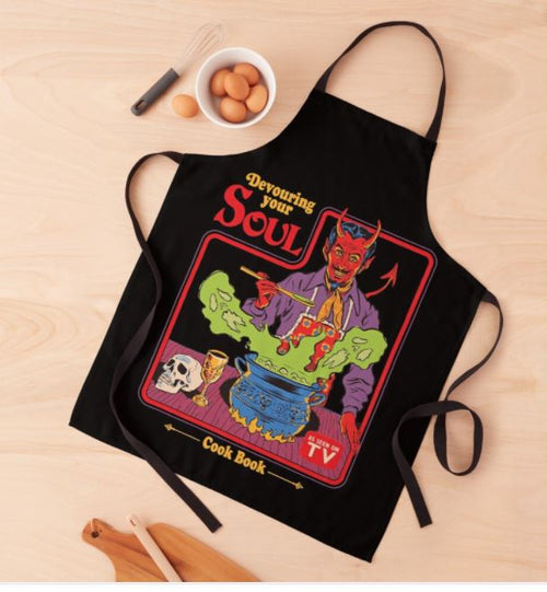 A Steven Rhodes Devouring Your Soul Apron from Blue Dog Posters sits on a wooden surface, showcasing a colorful design with a devil character cooking in a cauldron. The text reads "Devouring Your Soul" and "Cook Book." Nearby are a bowl of eggs, a wooden spoon, and a small blackboard. Plus, it's machine washable for easy care.