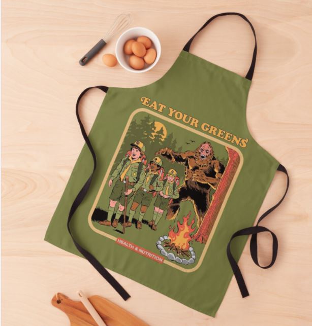 The Steven Rhodes Eat Your Greens Apron by Blue Dog Posters, adorned with a sublimation-printed design, is displayed on a light wooden surface. The illustration features four scouts gathered around a campfire near an apelike creature. The text "EAT YOUR GREENS" is prominent above them, with "HEALTH & NUTRITION" below. Placed nearby are a bowl of eggs and cooking utensils. This green apron is machine washable.
