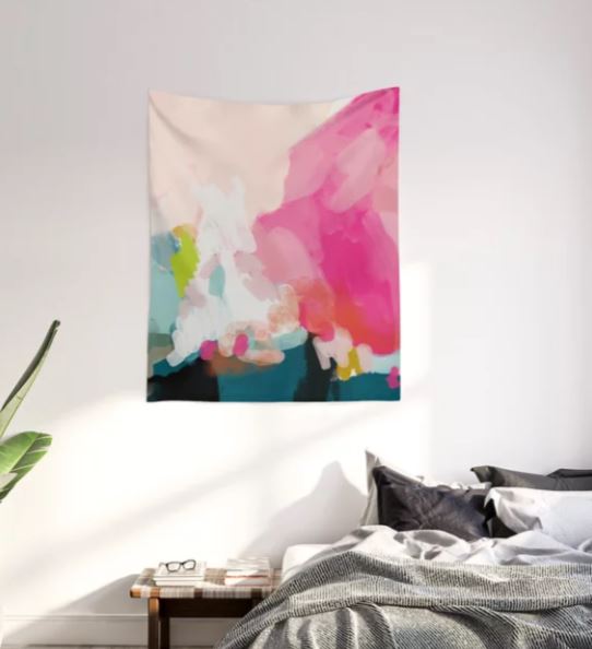 A Pink Sky Laluntricotee Art Textile Sml by Blue Dog Posters with splashes of pink, blue, and green hangs on a white wall above a bed. A small wooden nightstand next to the bed holds a pair of glasses and a book, and a green plant is placed to the left of the scene.