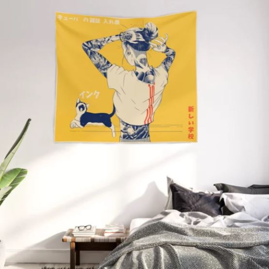 A yellow La Tinta Art Textile Sml from Blue Dog Posters hangs on a white wall above a bed with dark gray pillows and a white blanket. The tapestry, a striking piece of home decor, features an illustrated figure with tattoos and a raised arm, accompanied by Japanese text and a small black and white cat. A green plant is in the left corner.
