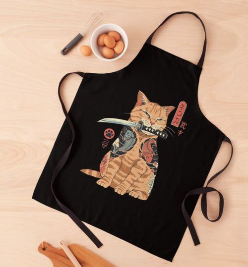 A black Catana Vincetrinidad Apron from Blue Dog Posters is laid on a wooden surface. The apron features an illustration of a cat holding a sword in its mouth, with intricately patterned designs and Japanese text. Nearby are cooking utensils and eggs. Plus, it's machine washable for easy care.