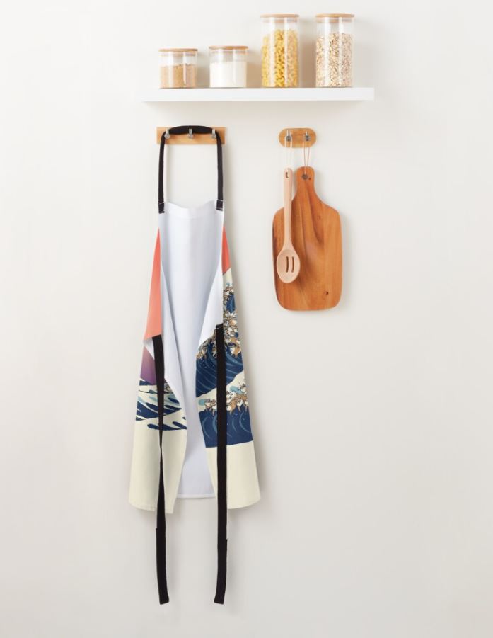 A kitchen apron from the Blue Dog Posters Homewares Collection, named The Great Wave of Shiba Inu Apron, hangs on a wooden hook. Above it is a white shelf holding glass jars filled with various food items. Next to the machine washable apron, two wooden cutting boards hold a spoon and spatula.