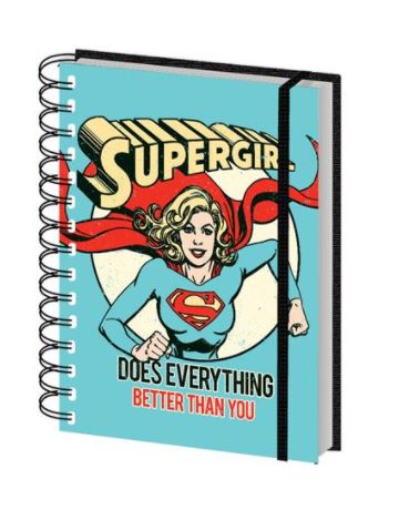 A high-quality hard cover spiral-bound notebook with a blue design featuring Supergirl flying with her fist forward. The text on the cover reads "SUPERGIRL" at the top and "DOES EVERYTHING BETTER THAN YOU" at the bottom in bold red and yellow letters. Introducing the DC Comics Supergirl A5 Spiral Notebook by Blue Dog Posters.