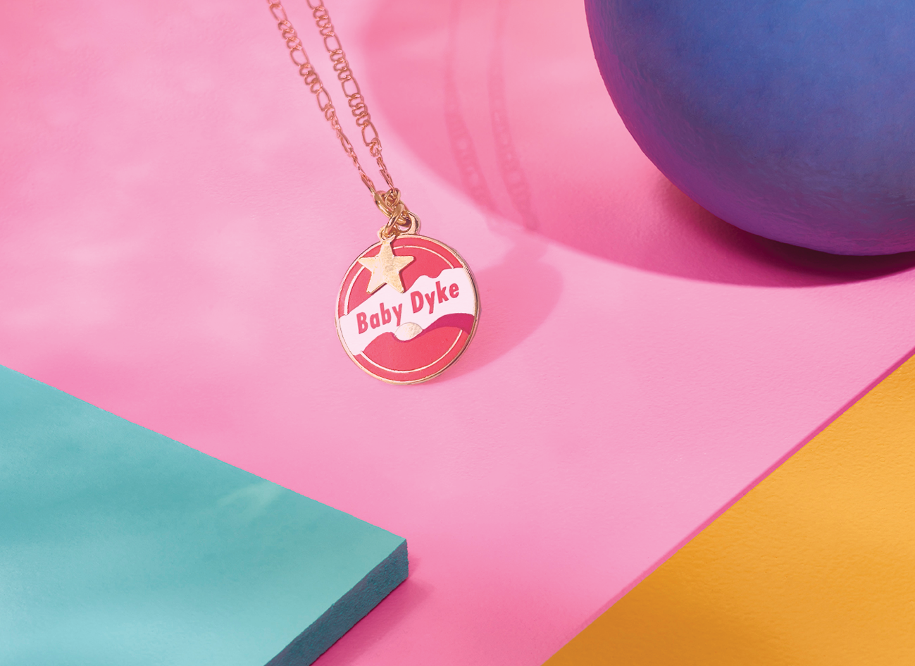 Introducing the Baby Dyke Necklace by Amy Blue, crafted by Blue Dog Posters. This unique piece of Queer Statement Jewellery features a 24k gold-plated chain with a gold-colored round pendant inscribed with "Baby Dyke" in striking red letters, complemented by white and red designs. It is beautifully showcased against a vibrant background with sections of pink, blue, and yellow.