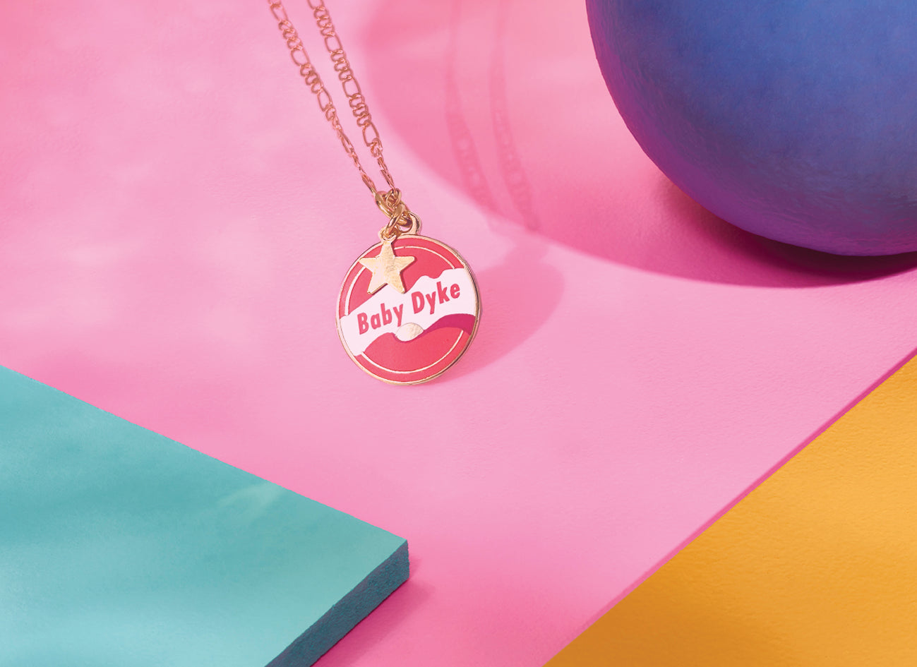 Introducing the Baby Dyke Necklace by Amy Blue, crafted by Blue Dog Posters. This unique piece of Queer Statement Jewellery features a 24k gold-plated chain with a gold-colored round pendant inscribed with "Baby Dyke" in striking red letters, complemented by white and red designs. It is beautifully showcased against a vibrant background with sections of pink, blue, and yellow.