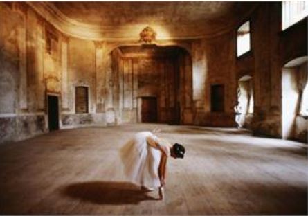 In the serene, vintage ambiance of a large, aged room with high ceilings, wooden floors, and partially curtained windows, a ballerina in a white tutu gracefully bends forward to touch her foot. This beautifully captures the essence of "Ballet Rehearsal" by ART 053—an ideal subject for an Art & Decor Poster.