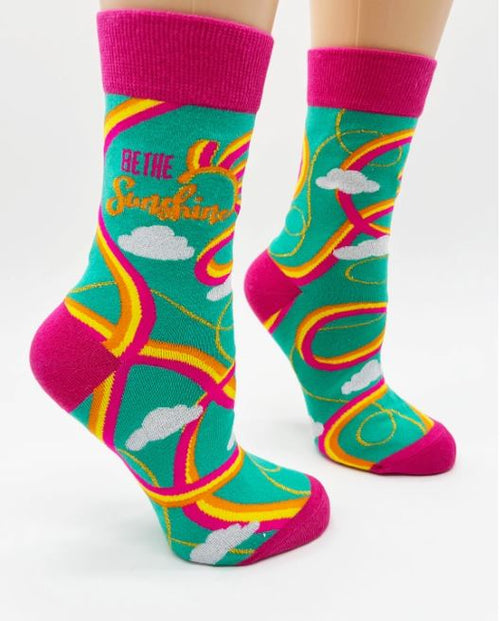 A pair of colorful Be The Sunshine Socks Ladies by Blue Dog Posters on a mannequin, showcasing a whimsical design with swirling patterns in pink, yellow, and orange against a turquoise background. These comfortable women's novelty crew socks feature hot pink cuffs, heels, and toes, with text that reads "BE THE Sunshine" accompanied by cloud motifs.