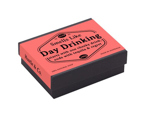 A rectangular black box with a pink label reads "Hustle and Co Soap - Day Drinking - starts with one cheeky drink, ends with tequila & regret." The label also includes "100% Natural Soap" and "Made in Australia." This plant-based Raspberry soap is suitable for all skin types and bears the Blue Dog Posters brand name.