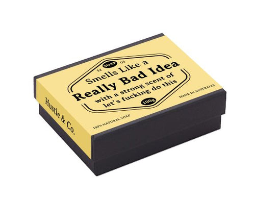 A rectangular black box featuring a yellow label that reads: "Hustle and Co Soap - Really Bad Idea" by Blue Dog Posters. The label displays the text "Smells Like a Really Bad Idea with a strong scent of let's fucking do this," and includes details such as "100g," "100% NATURAL SOAP," and "MADE IN AUSTRALIA." This luxurious mango soap is perfect for sensitive skin.
