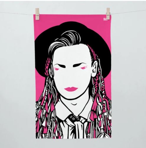 A stylized illustration of a person with a blank face, wearing a wide-brimmed hat and a shirt with a tie. The person's intricately braided hair features pink accents. The solid bright pink background, reminiscent of vibrant tea towels often seen in cozy kitchens, is held by two clothespins on a line. This design embodies the essence of the Rock and Pop Icons Tea Towel Boy George by Blue Dog Posters.