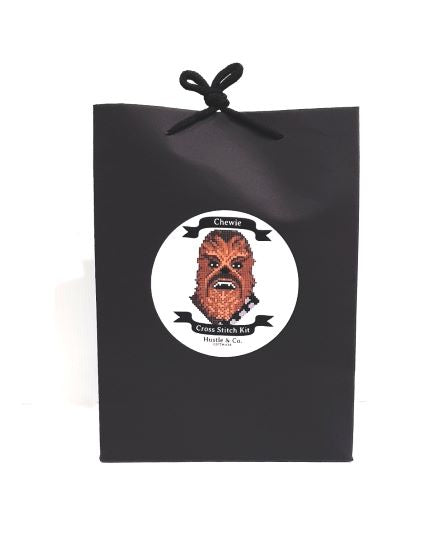 A black gift bag with a black ribbon handle. The bag features a circular label with an illustration of a character resembling Chewbacca from Star Wars, and the text reads "Hustle and Co. Cross Stitch - Chewie, Cool Gifts Collection, Blue Dog Posters.