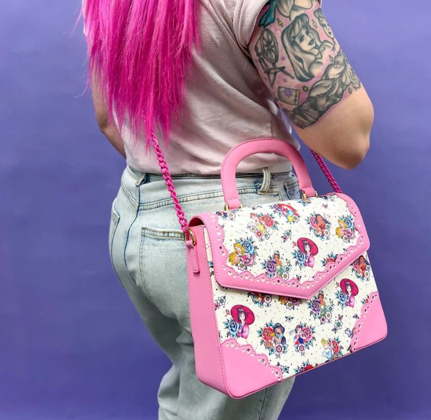 A person with long pink hair and vibrant arm tattoos is seen from behind, dressed in light-washed jeans and a pink top. They carry the Disney Princess Floral Tattoo Crossbody by Blue Dog Posters, a bag made from vegan leather with floral accents, cartoon character designs, pink highlights, and a chain strap set against a purple background.