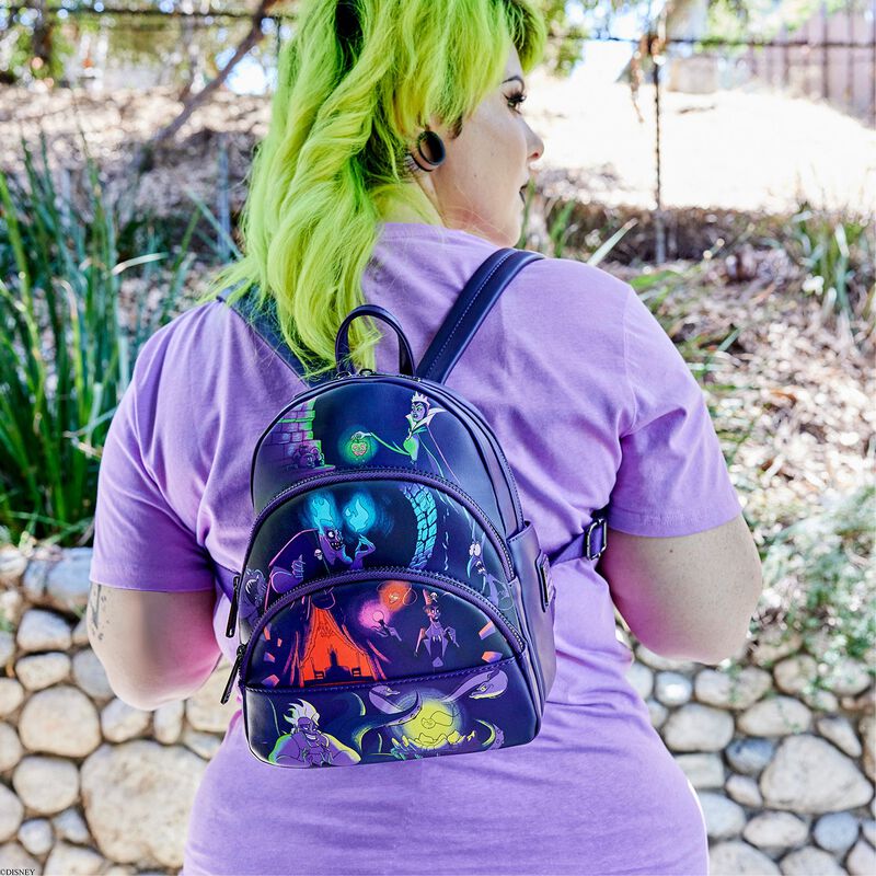 A person with bright green hair stands outdoors, wearing a purple T-shirt and a black Disney Villains Glow Mini Backpack by Blue Dog Posters, adorned with colorful, illustrated characters. The background features greenery and a stone wall.