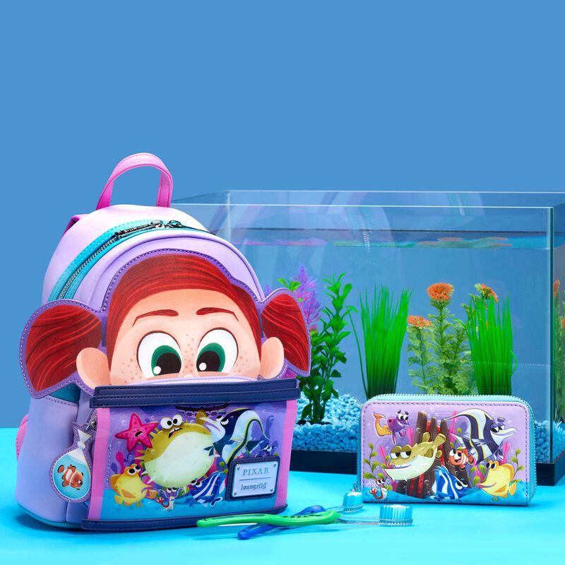 The Finding Nemo Darla Mini Backpack, featuring a cartoon girl design and ocean creatures by Blue Dog Posters, sits next to a matching wallet adorned with marine illustrations. In the background is an aquarium filled with colorful plants and gravel, while two toothbrushes and toothpaste lie in front.