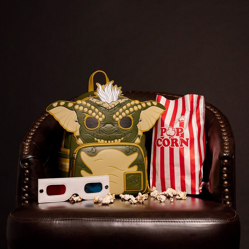 A Blue Dog Posters Gremlins Stripe Mini Backpack sits on a leather chair, accompanied by a striped red and white popcorn bag with some spilled popcorn and vintage 3D glasses. The backpack features a design that resembles a creature from the Gremlins movie.