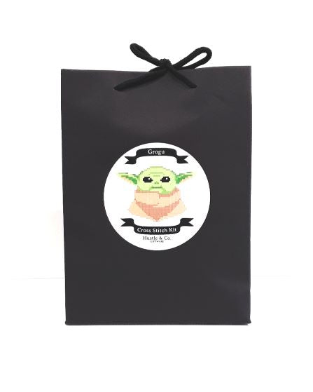 A black gift bag with a tied handle features a circular label showcasing a cross-stitched Grogu from "The Mandalorian." The label reads "Hustle and Co. Cross Stitch - Grogu, Blue Dog Posters." Part of the Cool Gifts Collection, this kit includes an embroidery hoop for your stitching delight.