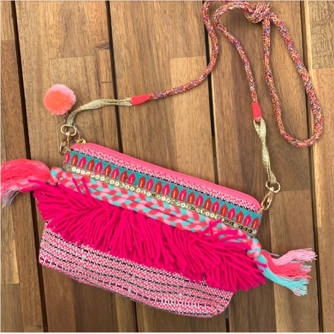 The Hot Pink Hand Woven Tassels Bag by Blue Dog Posters, featuring a colorful, bohemian-style design with vibrant patterns in pink, teal, and gold, lies on a wooden surface. The bag boasts decorative fringes and tassels, along with a pink pom-pom attached to one end of the removable long braided shoulder strap.