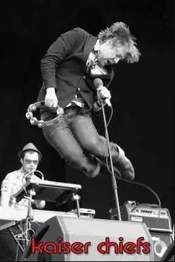Black and white image of a band performing on stage. The lead singer, mid-jump, holds a tambourine and a microphone. Another band member is playing a keyboard in the background. The text "Kaiser Chiefs" appears at the bottom in red font, making this MUS 207 rock poster a standout addition to any music collection.