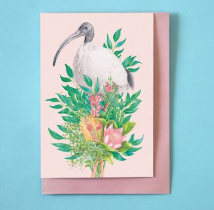 The Kayla Reay Ibis Greeting Card from Blue Dog Posters features a detailed illustration of a white ibis with a long, curved beak standing among vibrant flowers and green foliage on a pale pink background. Made with quality materials, it comes in a matching pale pink envelope set against a light blue surface.