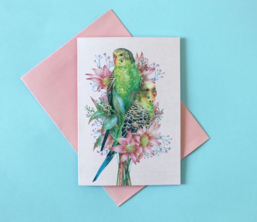The Kayla Reay Budgies Greeting Card from Blue Dog Posters features two beautifully illustrated green and yellow parakeets perched on pink flowers, set against a light blue background. The vibrant colors really make the design stand out. This stunning card comes with a matching pink envelope.