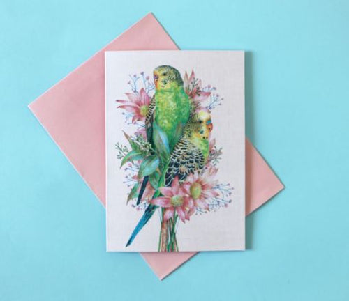 The Kayla Reay Budgies Greeting Card from Blue Dog Posters features two beautifully illustrated green and yellow parakeets perched on pink flowers, set against a light blue background. The vibrant colors really make the design stand out. This stunning card comes with a matching pink envelope.