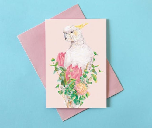 The **Kayla Reay Cockatoo Greeting Card** from **Blue Dog Posters** features a detailed illustration of a white cockatoo with a yellow crest perched among vibrant pink and orange flowers and green leaves. Made with quality materials, the card is placed on top of a pink envelope against a light blue background.