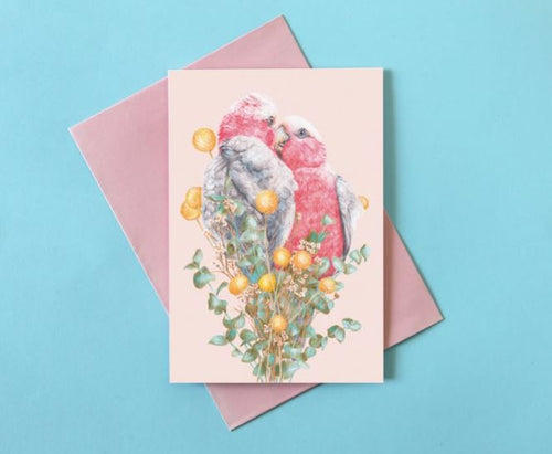 A Kayla Reay Galah Greeting Card from our Blue Dog Posters collection features an illustration of two pink and grey birds perched closely together on a branch adorned with yellow flowers and green leaves. Crafted from quality materials, the card is placed on a slightly larger pink envelope, set against a light blue background.