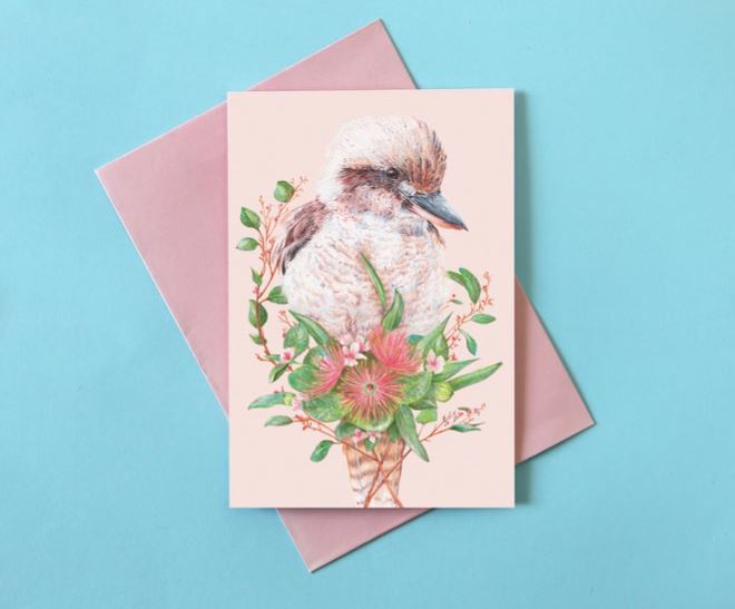 The Kayla Reay Kookaburra Greeting Card from Blue Dog Posters, featuring an illustration of a kookaburra surrounded by vibrant flowers and greenery, placed atop a pink envelope on a blue background. The bird and floral design are detailed with soft colors, using quality materials to create an elegant and cheerful image.