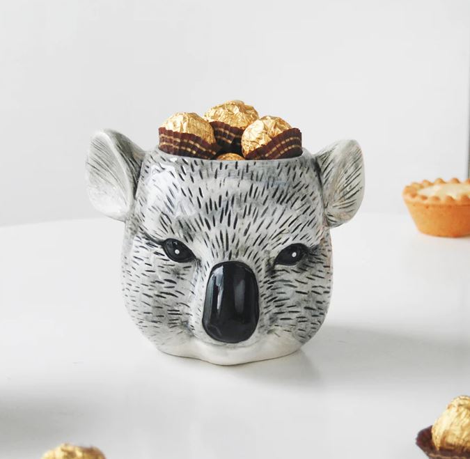 A Ceramic Planter - Koala by Blue Dog Posters, filled with wrapped chocolate candies, showcases detailed black and grey markings to resemble a koala's face. Placed against a blurred white surface with another dessert mildly visible to the side, this versatile planter could also double as an adorable mug for your favorite beverages.