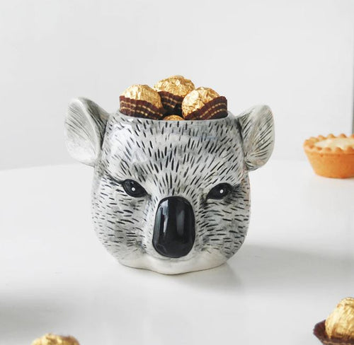 A Ceramic Planter - Koala by Blue Dog Posters, filled with wrapped chocolate candies, showcases detailed black and grey markings to resemble a koala's face. Placed against a blurred white surface with another dessert mildly visible to the side, this versatile planter could also double as an adorable mug for your favorite beverages.