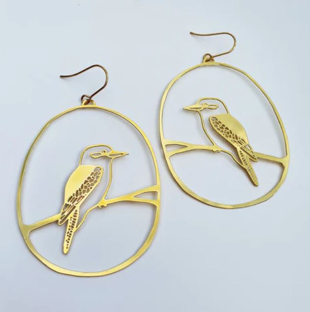 The Denz Kookaburra Dangles Gold Earrings from Blue Dog Posters feature a pair of statement earrings with an open oval frame and a detailed depiction of a Kookaburra perched on a branch in the center. Crafted from recycled plated stainless steel, these delicate and intricate earrings showcase the bird's feathers and the texture of the branch, hanging elegantly from simple gold hooks.