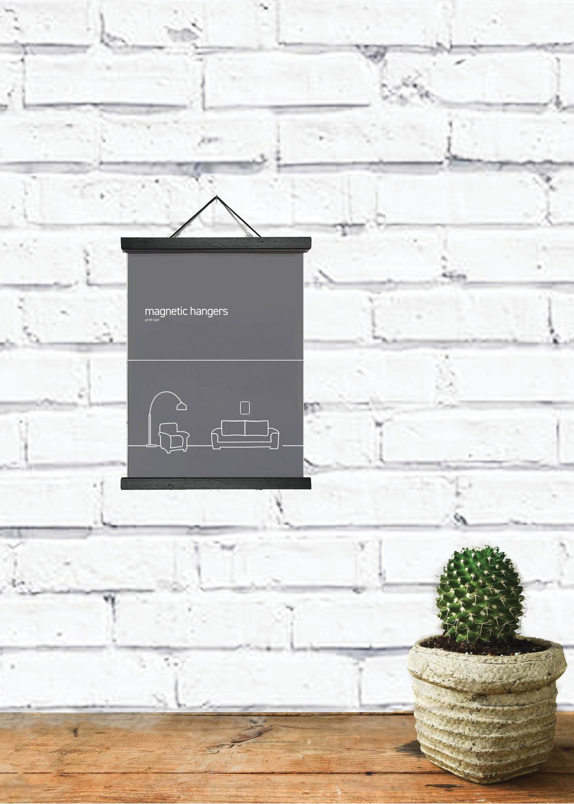 A minimalist room decor setup features a small green cactus in a woven pot on a wooden surface. A black and white poster is elegantly displayed with Blue Dog Posters' Magnetic Timber Hanger 15cm Black, hanging on a white brick wall in the background.