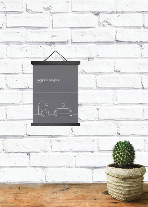 A minimalist room decor setup features a small green cactus in a woven pot on a wooden surface. A black and white poster is elegantly displayed with Blue Dog Posters' Magnetic Timber Hanger 15cm Black, hanging on a white brick wall in the background.