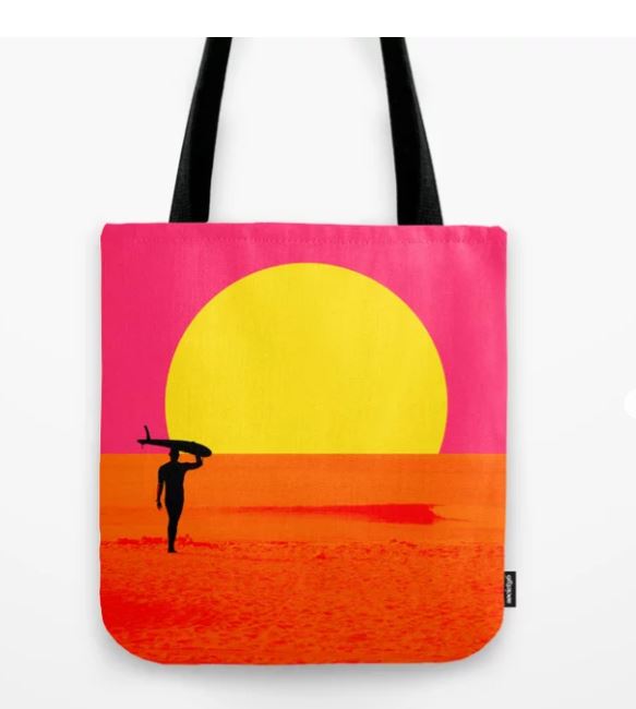 A **Tote Bag 40x40cm Malibu Surfers** featuring an artistic design of a surfer carrying a surfboard on a beach. The silhouette of the surfer is set against a vibrant pink and orange sky with a large yellow sun setting in the background. Made from quality blended fabric, the bag has black straps and is sublimation printed. Available from **Blue Dog Posters**.
