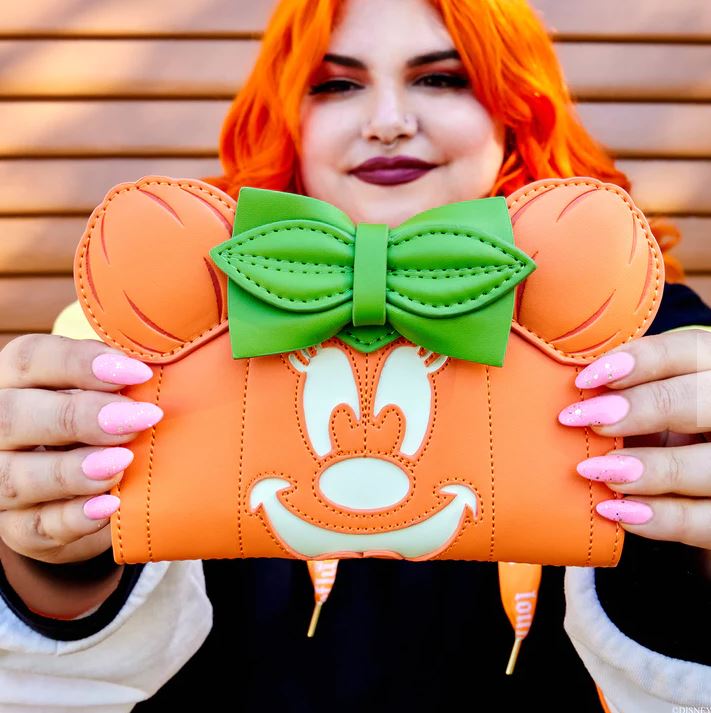 A person with vibrant orange hair holds up the Disney Minnie Pumpkin Face Flap Purse by Blue Dog Posters, featuring a cartoon mouse face with a giant green bow. The individual, showcasing long pink nails and a broad smile, exudes joy. This Halloween accessory pops against the wooden wall backdrop, capturing festive charm in every detail.