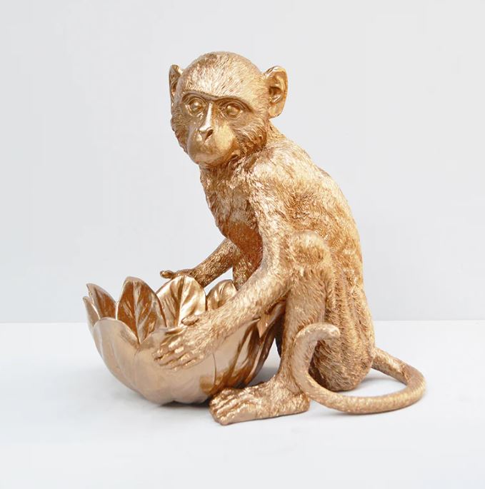 A gold-colored statue of a monkey sits with its front paws resting on a large, lotus-shaped resin bowl. The monkey's detailed fur texture and the ornate design of the Monkey Bowl Gold from Blue Dog Posters stand out against a simple, white background, making it perfect for an elegant snack display.