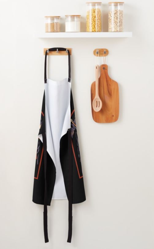 A kitchen scene featuring a white shelf with three jars of dried goods on top. Below the shelf, a Steven Rhodes Morning Ritual Apron by Blue Dog Posters, which is black and machine washable with a white lining, hangs on a hook next to a wooden cutting board and spoon set also hung on the wall. This display is part of our stylish Homewares Collection.