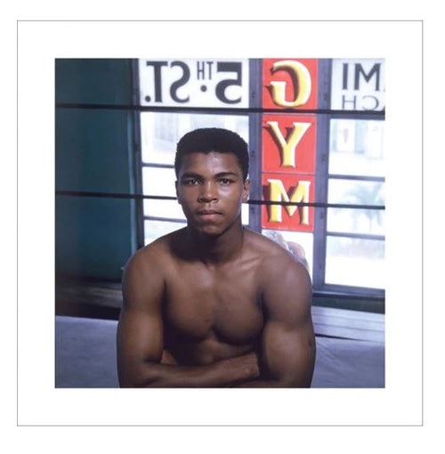 A young man with a muscular build sits shirtless in a gym, his face reflecting deep thought. Behind him, windows emblazoned with "Muhammad Ali Gym" in bold red letters from Blue Dog Posters and other signage are visible.