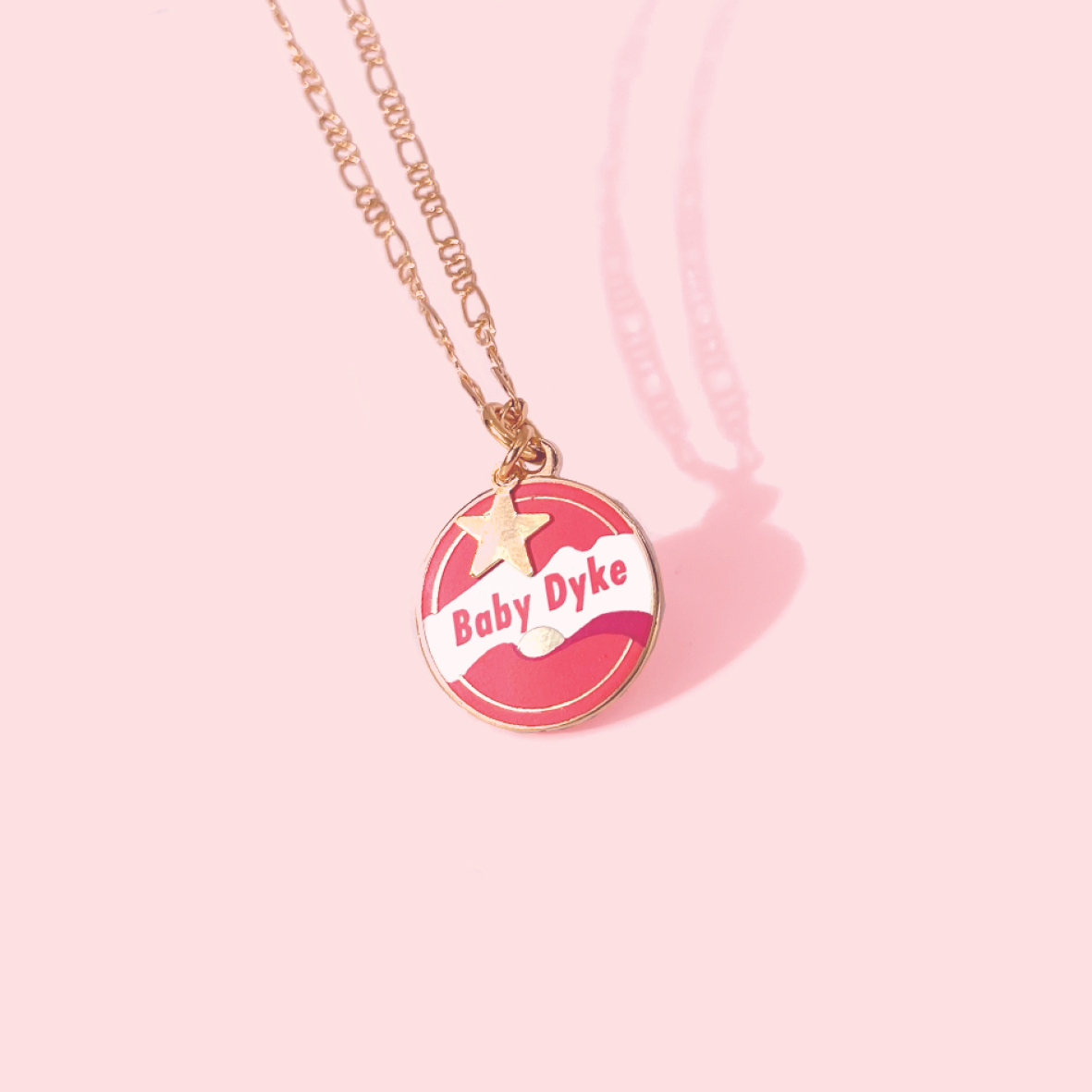 The Baby Dyke Necklace by Amy Blue, offered by Blue Dog Posters, is a stunning gold necklace featuring a circular 24k gold plated pendant with a pink and white design, showcasing a star and the text "Baby Dyke." Perfectly set against a pink background, this piece makes an ideal piece of queer statement jewelry.