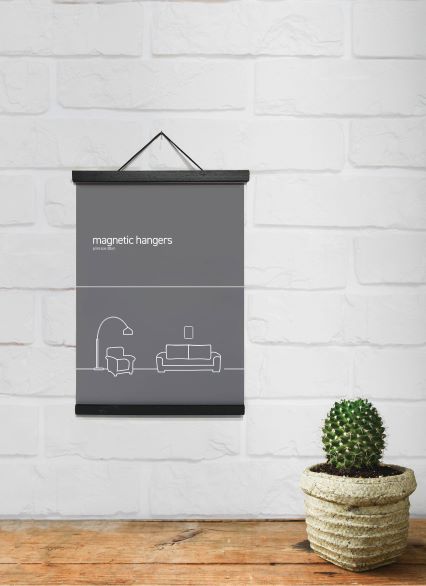 A minimalist poster with the text "Magnetic Timber Hanger 21.5cm Black" and an illustration of a lamp, couch, and picture frame on a dark gray background hangs on a white brick wall. A small potted cactus sits on a wooden surface below. The black finish of the magnetic hanging strips adds an elegant touch, featuring the Blue Dog Posters branding.