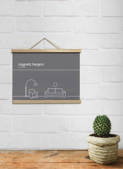 A minimalist room with a light gray brick wall features a hanging poster labeled "Magnetic Timber Hanger 43cm Natural" by Blue Dog Posters above a small illustration of a living space. Below, a potted cactus sits on a wooden floor, adding a touch of greenery to the scene.