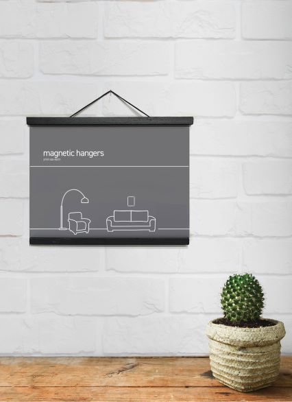 A minimalist poster by Blue Dog Posters, titled "Magnetic Timber Hanger 43cm Black," is displayed in a sleek black frame against a white brick wall. Below, a small green cactus in a beige pot rests on a wooden surface. The poster, ideal for enthusiasts of hanging posters, showcases simple line drawings of a lamp, couch, and picture frame.
