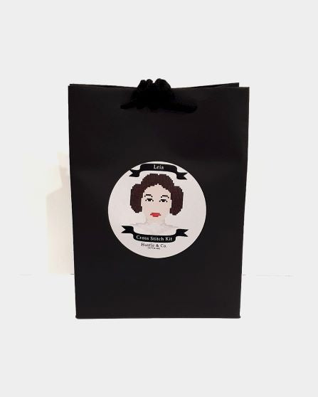 An upright, black gift bag with black handles features a circular illustration of a woman with brown hair styled in buns on each side of her head, likely a cross-stitch design. Below the illustration, the text reads "Hustle and Co. Cross Stitch - Leia," part of the Cool Gifts Collection by Blue Dog Posters.