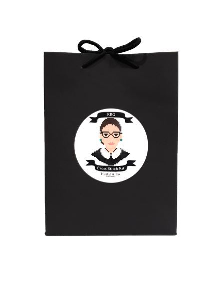 A black gift bag with a black ribbon handle and a round white label in the center. The label features a pixelated cross-stitch image of a person with glasses and the text "Hustle and Co. Cross Stitch - RBG" from Blue Dog Posters, part of the Cool Gifts Collection.