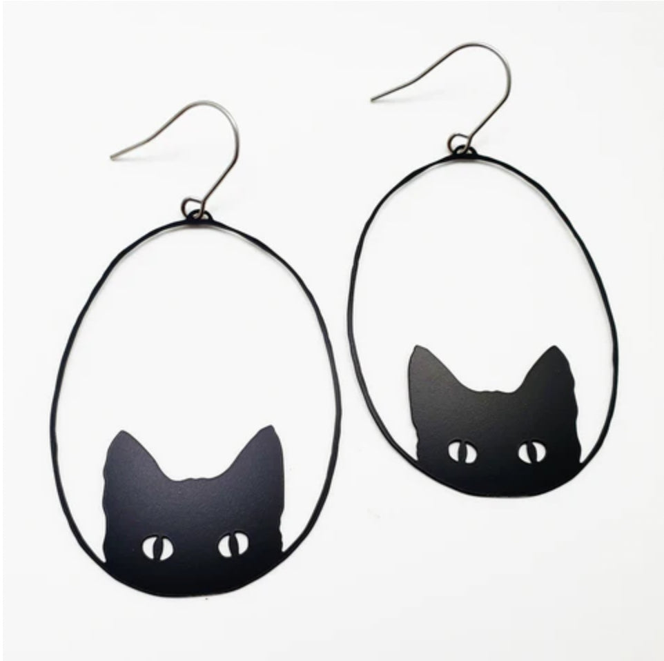The Denz Cat Black Earrings by Blue Dog Posters are black cat-themed and handmade in Noosa. They showcase an outline of a cat’s head with pointed ears and white eyes on a simple, minimalist white background. Crafted from recycled stainless steel, these lightweight statement earrings are framed in oval wire hoops with hooks for wearing.